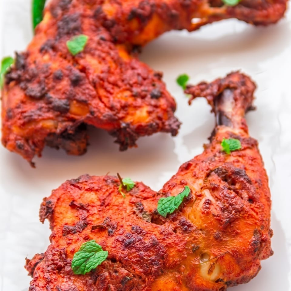 Two red tandoori chicken legs garnished with mint leaves, lime, and onion slices.